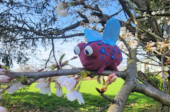 A clay frog model with, purple skin, blue spots, and wings sits in a flowering tree.