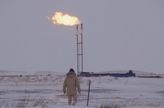 a person in a large white puffer jacket stands in a snowy field on a overcast day. in the distance, two tall pipes emit a flame blowing in the wind