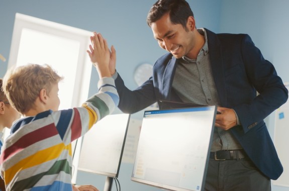 The image shows an elementary school computer science classroom that is bright and colorful, with posters and artwork on the walls. A male young student is receiving a high-five from his AAIP male teacher, who is smiling with encouragement.