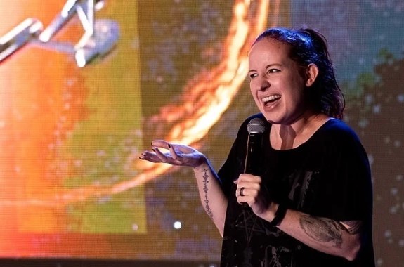 an astrophysicist with really cool tattoos on her arm and with her hair tied in a ponytail smiles and talks into a mic with her hand propped out in gesture. behind her is a screen display of a futuristic space ship, from the star trek series, flying towards a planet