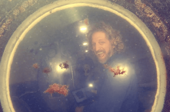 a man smiles through a circular window of an underwater lodge. the exterior of the lodge is slightly crusty with ocean debris and you can see floating debris in the water