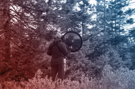 a woman holds a large dome-shaped recording device in a forest. the image is tinted red and blue