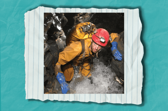 A man wearing a yellow jumpsuit and a helmet stands inside a cave full of water.
