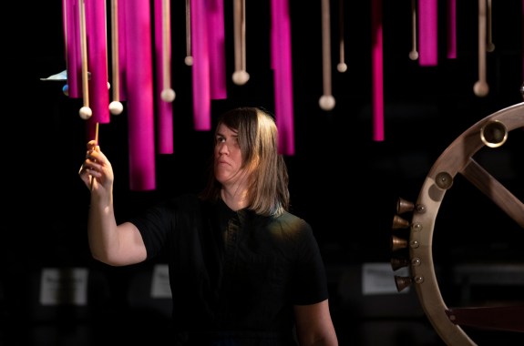 A white woman in a black shirt in a dark room raises a mallet among various brown mallets and hot pink pipes hanging down around her.