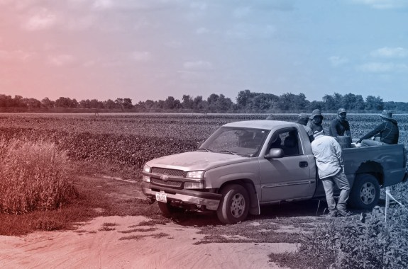farmworkers sit and stand in the back of a silver pickup truck in the middle of a farm field. the image has a red and blue filter