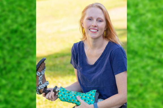 A white woman sitting outside in the sun smiles while holding a prosthetic leg with a sandal on the foot
