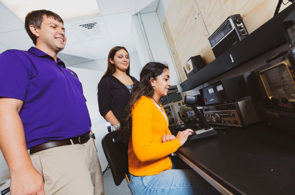 A man and woman stand behind a young woman sitting at a desk operating a ham radio.