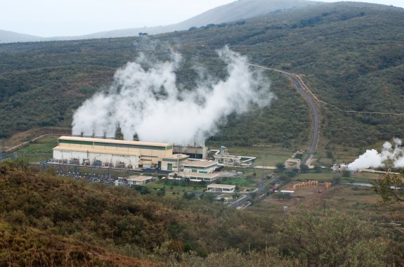 Plumes of steam drift up from a line of cooling towers behind a long, beige building. The structures are set in green, arid valley between two hills.