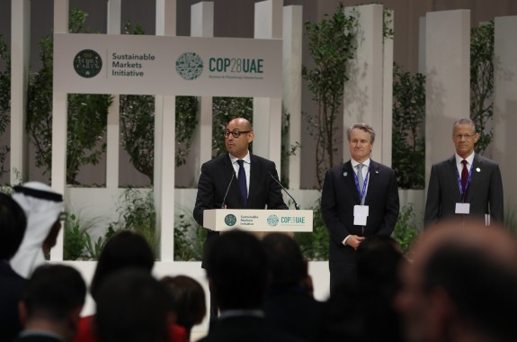 A man stands at a podium speaking to a crowd. Behind him is a sign that says "COP28UAE"