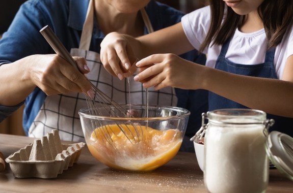 Close up of hands as grandkid helps grandma cook, cracking eggs and whisking them in glass bowl.