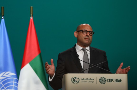 A man in a suit gives a speech, looking tired against a green background