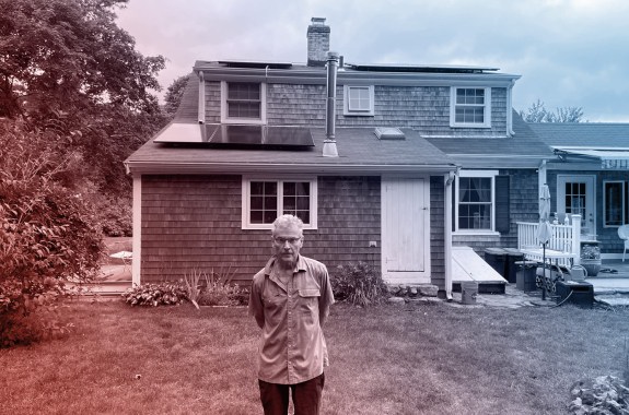 A man stands on the lawn in front of a house with 3 black solar panels on the roof.