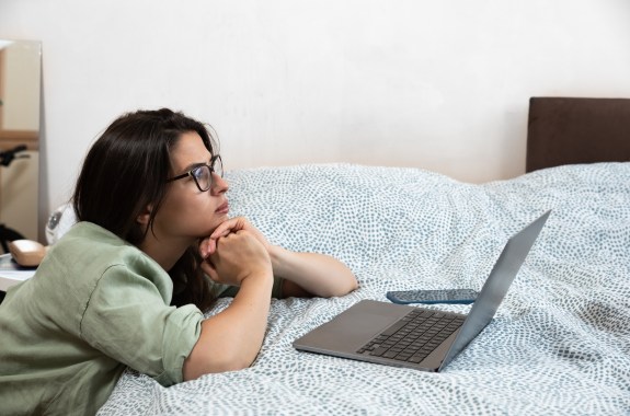 A young woman wearing glasses looks at her laptop on her bed.