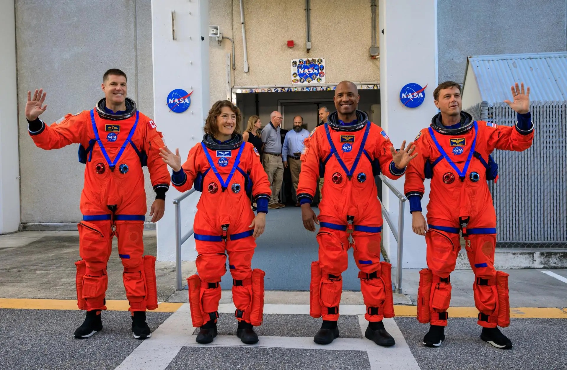 Four NASA astronauts standing in bright orange space suits waving at the press.