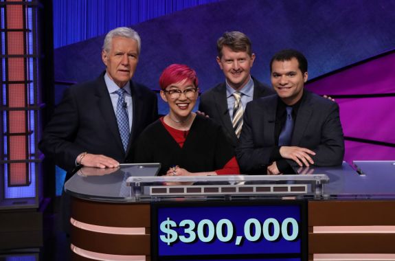 Four smiling people standing at a Jeopardy! games desk with "$300,000" on the screen.
