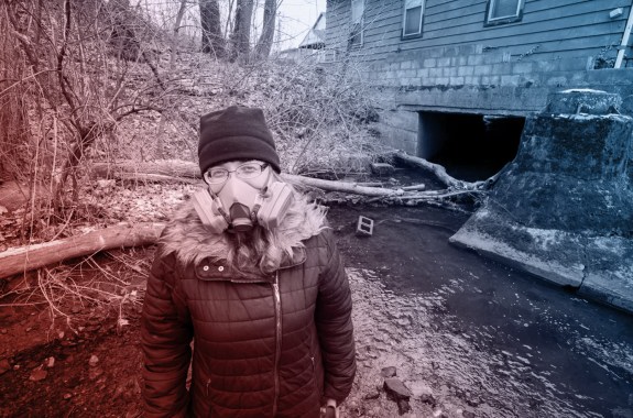 A woman wearing a gas mask over her mouth and nose standing in front of a muddy, polluted body of water.