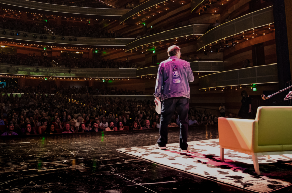 Ira Flatow stands on a stage addressing a live audience in a large auditorium. He faces away from the camera.