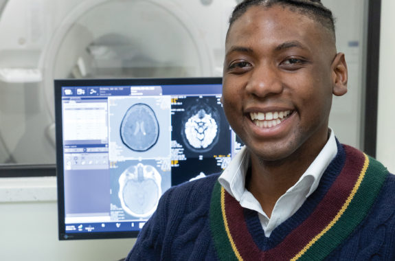 A Black man in a blue sweater and white collared shirt smiles at the camera. Behind him is an MRI machine and brain scans on a monitor.
