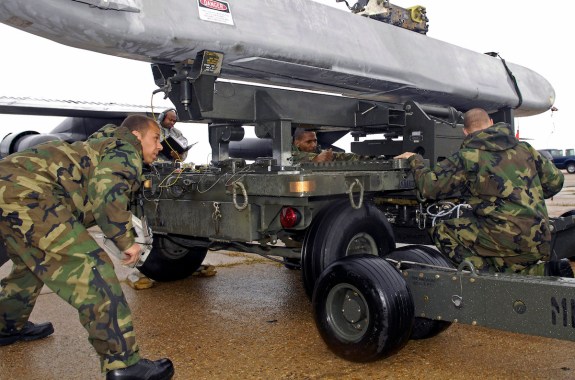 Four men in camo are crouched around the base of a large platform holding a long, gray missile-like object.