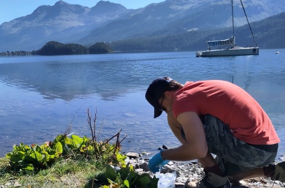 A man wearing gloves crouches by a lake, about to collect samples of the water,