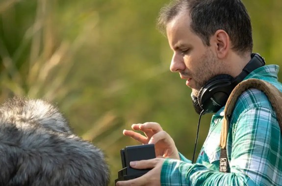 A side view of a man in a green plaid shirt with headphones around his neck and a large fuzzy mic cover handling recording equipment against a blurry green background.