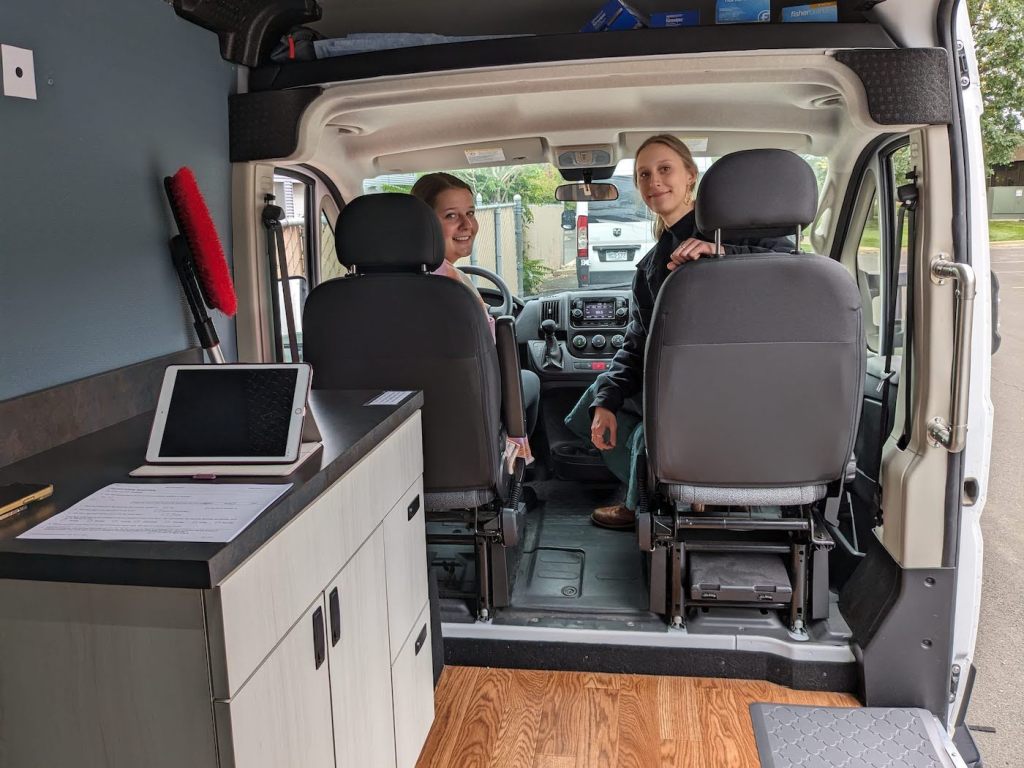 Two women sit in the driver and passenger's seat of a van, which has a small medical lab in the back.