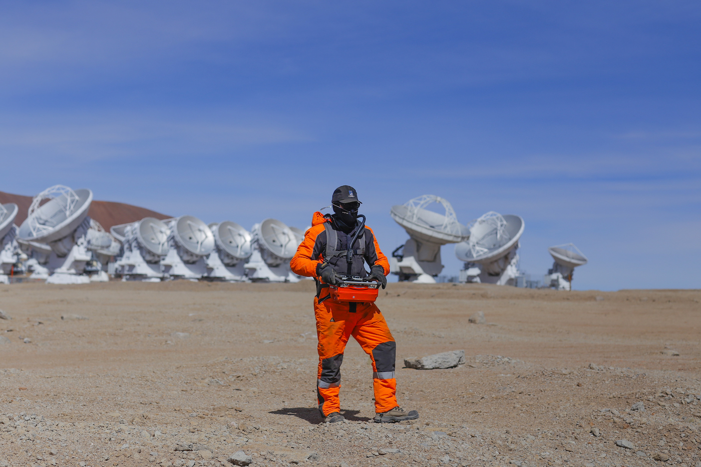 A person wearing an orange jumpsuit standing on rocky terrain. In the background are a bunch of sattelites.