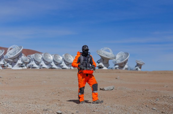 A person wearing an orange jumpsuit standing on rocky terrain. In the background are a bunch of sattelites.