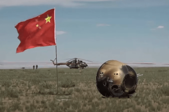 a metal capsule next to a planted Chinese flag on a grassy plain.