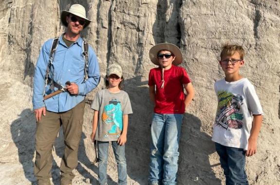 On the far left, an adult man wearing a sun hat. On the right, three young boys. They are all standing in front of a large rock wall outdoors.