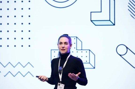 a white woman wearing a black turtleneck wearing a lanyard and a necklace mic gesturing with her hands speaking on a stage with abstract graphics behind her