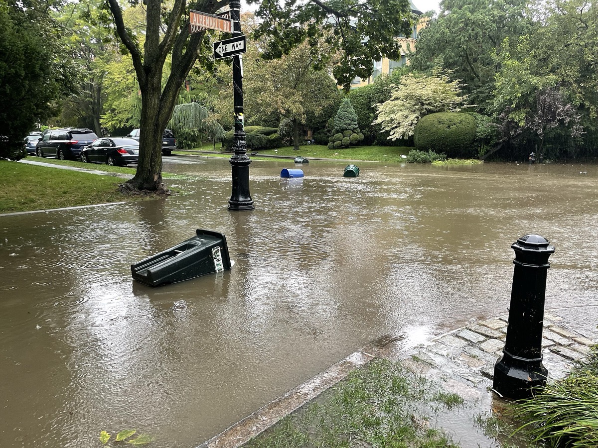 A residential neighborhood's streets flood with dirty brown water. Trash cans lie toppled in the water as it continues to rain.