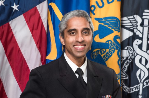 A man wearing an official US uniform in front of 4 flags, including the American flag.