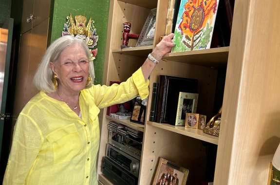 An older woman looking happy and healthy, reaching up at a painting she made on a shelf.