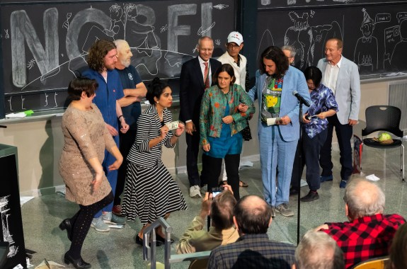 A woman holds a dollar bill on a stage with several others in expressive outfits stand by her.