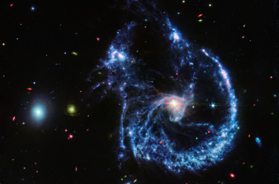 A pair of interacting galaxies. The larger of the two galaxies is slightly right of center, and is composed of a bright, white center and a ring of blue, gaseous filaments. The center of this galaxy shows Webb’s eight-pronged diffraction pattern. There are three filaments of gas and dust moving from the ring toward the center. At the top left of the ring is a noticeable gap, bordered by two large, blue pockets of dust and gas. The smaller galaxy is made of hazy, light blue gas and dust. Many red, green, blue, and yellow galaxies are spread throughout, with some being hazier in composition and others having more defined spiral patterns.