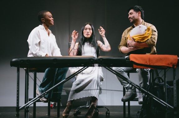 Three people sit on a stage, performing, The woman in the center is hooked up to a bunch of wires, and the man to the right is holding something like a baby