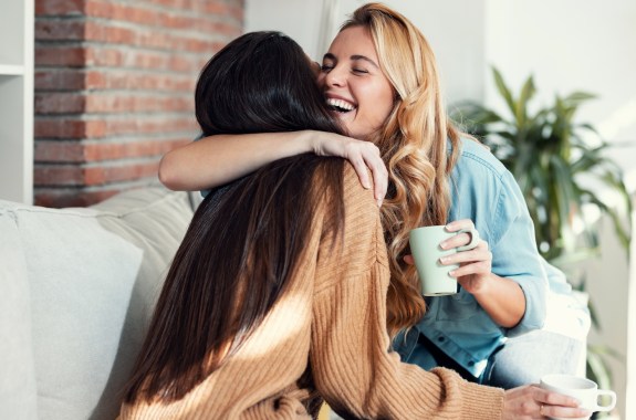 Shot of two lovely smiling women hugging each other while talking sitting on couch in the living room at home.