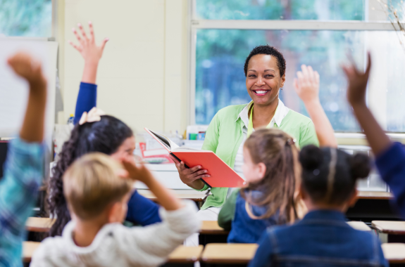 A teacher holding a book in a classroom with students raising their hands.
