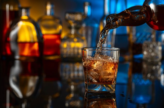 Whiskey is poured from a bottle into a glass with ice. Glass of whiskey on a black reflective background. In the background bottles with various alcoholic drinks.