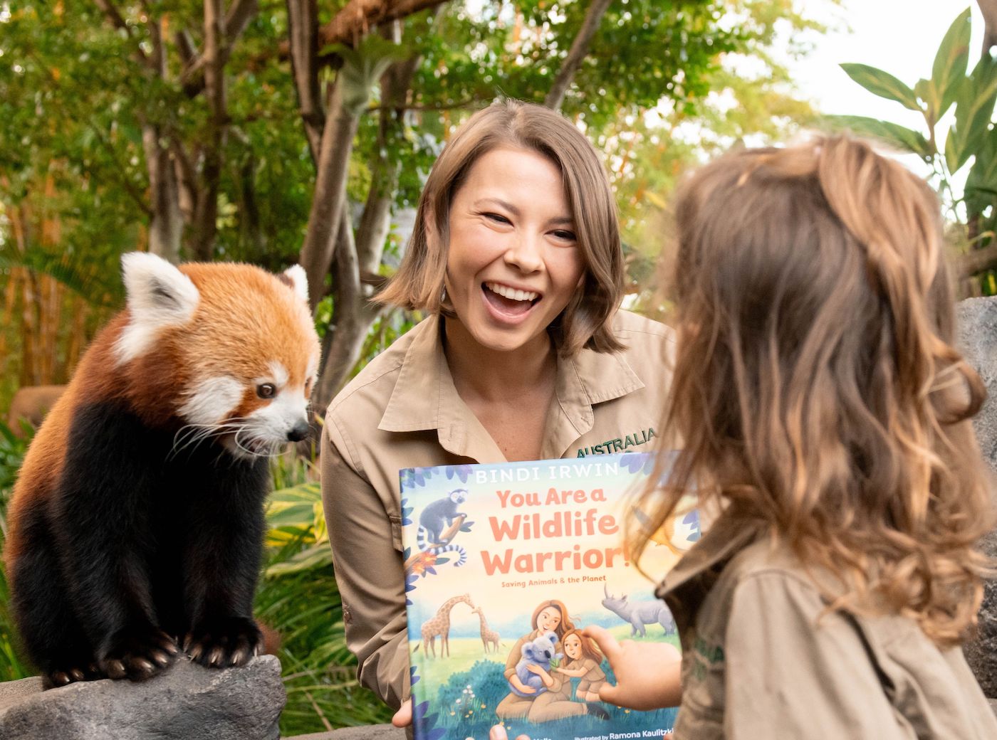 A happy woman holding a book, showing it to a young child. She sits next to a red panda.