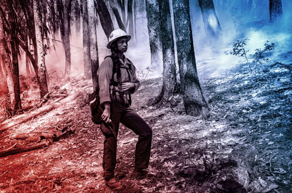 A woman wearing a construction helmet stands holding a radio in a smoky wooded area.