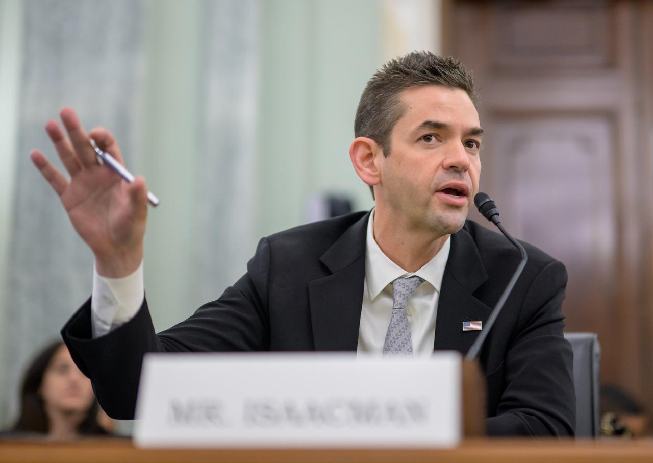 A man speaking into a microphone at a desk. A name card in front of him says "Mr. Isaacman"