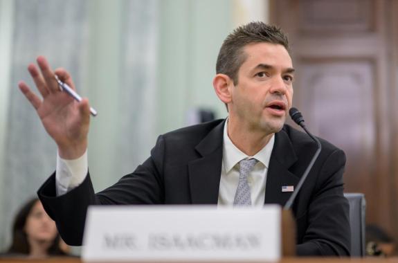 A man speaking into a microphone at a desk. A name card in front of him says "Mr. Isaacman"