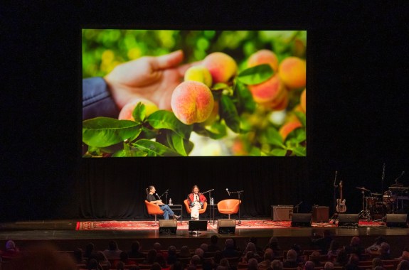 Two people sitting on a large stage with a picture of peaches projected behind them.