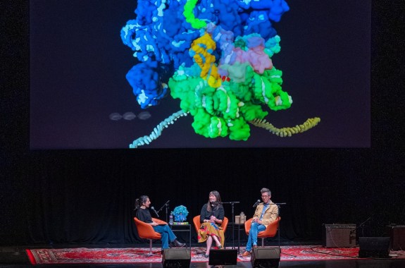 A large colorful 3-d model of a complex biological subject is projected above three people speaking on a stage.