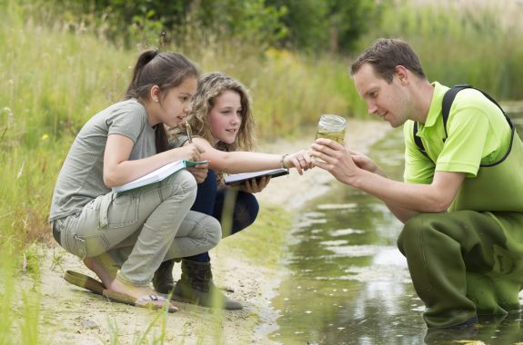 Learners and an educator outdoors by the side of a pond. The educator is holding a glass jar of pond water as the students record observations in their notebooks. Credit: Shutterstock