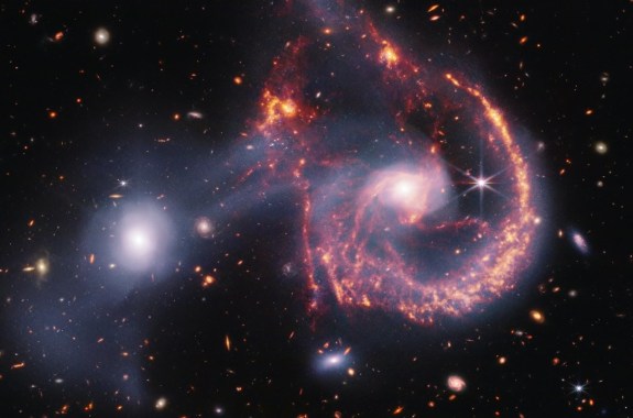 A pair of interacting galaxies. The larger of the two galaxies is slightly right of center, and composed of a hazy, bright, white center and a ring of gaseous filaments, which are different shades of red and orange. Toward the bottom left and bottom right of the ring are filaments of gas spiraling inward toward the core. At the top left of the ring is a noticeable gap, bordered by two large, orange pockets of dust and gas. The smaller galaxy to its left is made of hazy white gas and dust, which becomes more diffuse farther away from its center. To this galaxy’s bottom left, there is a smaller, more diffuse gas cloud that wafts outward toward the edges. Many red, orange, and white background galaxies are spread throughout, with some hazier in composition and others having more defined spiral patterns.