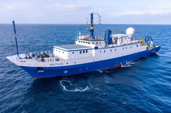 A white and blue research vessel, the E/V Nautilus, floats on a bright blue sea.