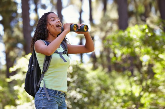 Happy girl, binoculars and nature with backpack for sightseeing, explore or outdoor vision. Young female person, child or teenager enjoying sight, adventure or bird watching and search in forest